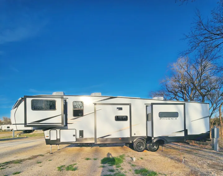 A large white Phoenix fifth-wheel RV rental with slide-outs extended, parked on a gravel lot under a clear blue sky.. phoenix 334FL 2026