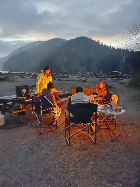 Friends enjoy a cozy evening at an RV rental campsite. Warm light from a heater illuminates their gathering under misty mountains.. Volkswagen T3 Westfalia 1985