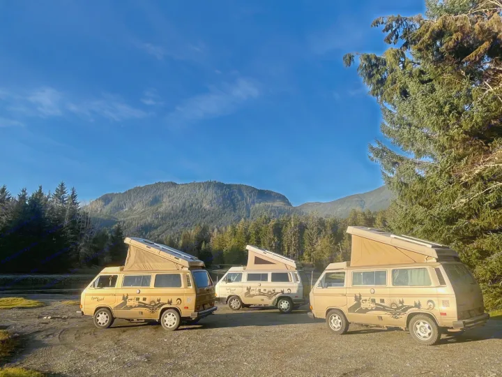 Three vintage tan "Open Road" rv rental campervans with pop-tops, parked amidst mountains & forest under a clear blue sky.. Volkswagen Westfalia 1986
