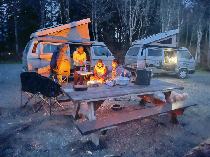 Friends enjoy a cozy evening at a campsite with two pop-top RV rental vans and a dog. Warm light illuminates the scene.. Volkswagen Westfalia 1989