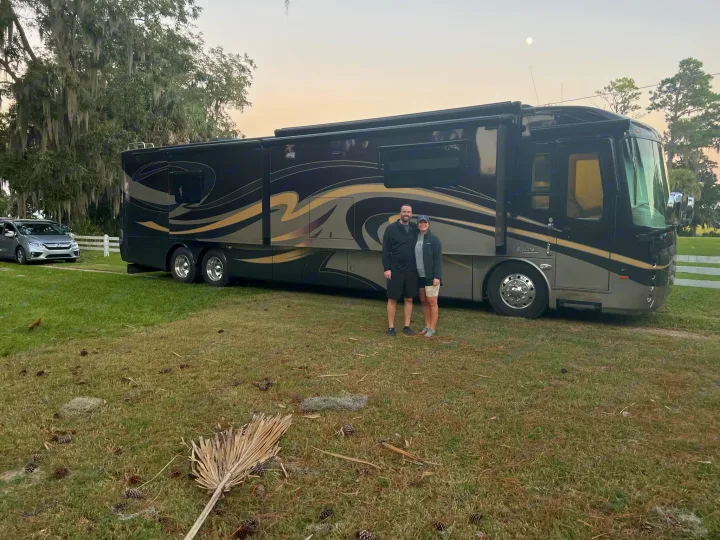 A couple stands with their elegant black and gold rv rental, parked on grass with a silver car, mossy trees, and moon overhead.. Entegra Coach Aspire 2015