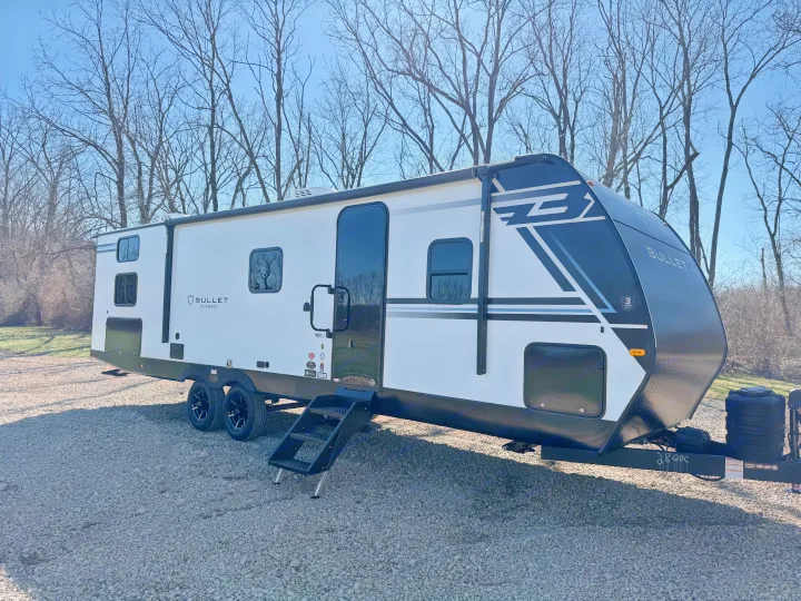 White & black Bullet Classic travel trailer RV rental parked on a gravel lot under a clear blue sky, surrounded by bare trees.. Keystone Bullet 2026