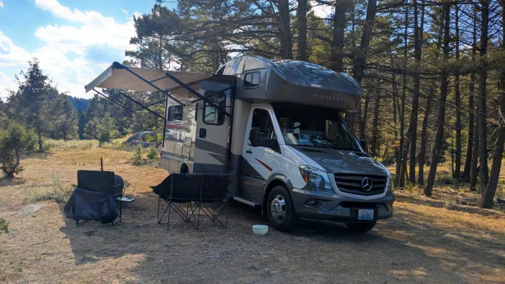 This Winnebago rv rental with an extended awning and chairs sits in a sunny, forested campsite, ready for outdoor adventure.. Winnebago View 2017