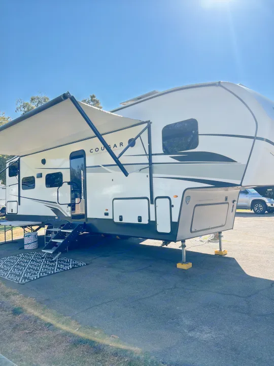 This white Cougar fifth-wheel RV rental is set up at a sunny campsite with its awning extended and an outdoor rug.. Keystone RV Cougar 2023