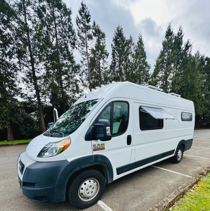 White Ram ProMaster RV rental van with custom features, including an open side window, parked by tall trees on a cloudy day.. Custom Camper 2021