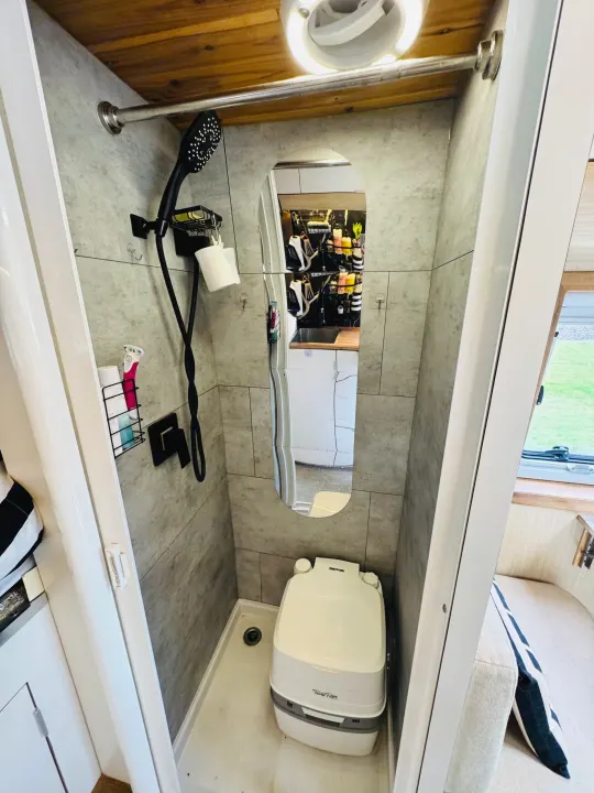 This rv rental's compact wet bath features a portable toilet, shower, and a mirror reflecting the kitchenette area.. Custom Camper 2021