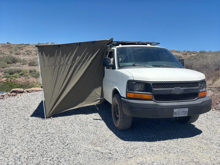 This white RV rental camper van with an olive green awning is set up by a pond for adventure in a sunny, gravelly landscape.. Chevrolet Express 1500 AWD 2007