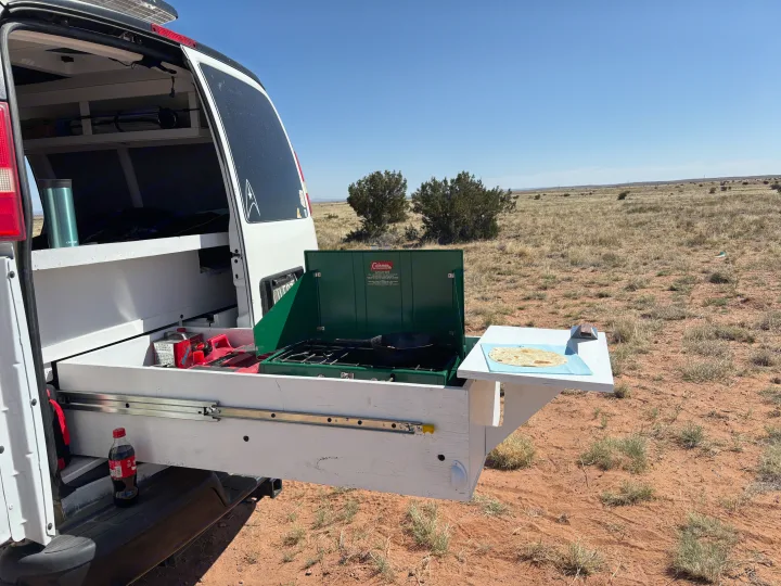 RV rental outdoor kitchen: A pull-out stove from a camper van cooking in an arid landscape with tortillas on a side table.. Chevrolet Express 1500 AWD 2007