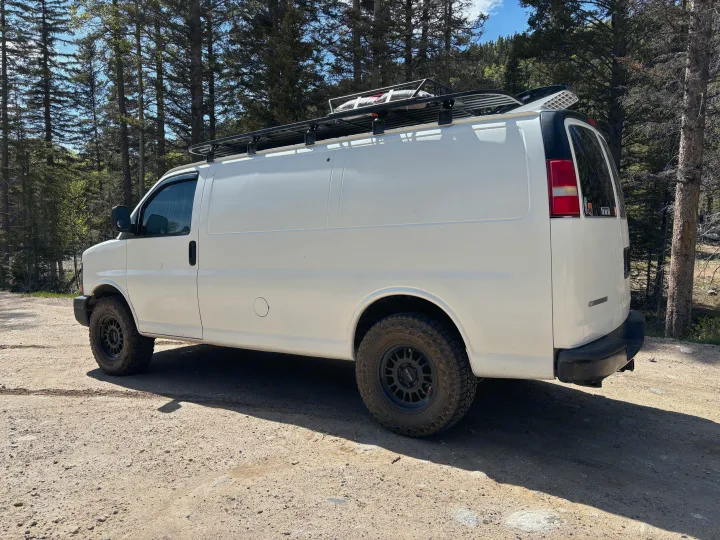 White camper van, equipped with a roof rack and off-road tires, parked on a forest dirt road. Ideal for an RV rental adventure.. Chevrolet Express 1500 AWD 2007