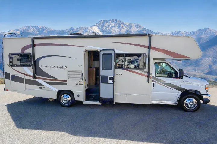 RV rental: Beige Coachmen Leprechaun motorhome with open door, parked against majestic snow-capped mountains. Ideal for scenic trips.. Coachman Leprachaun 2020