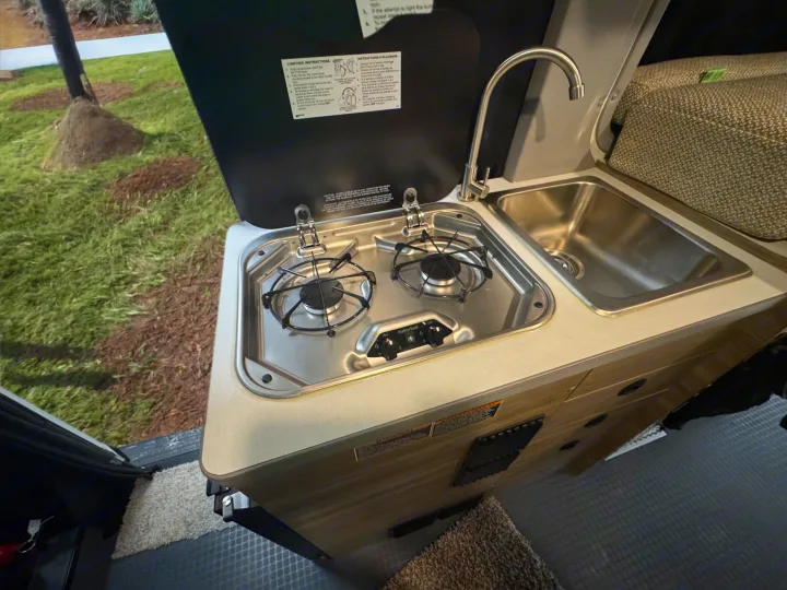 A compact kitchenette in an RV rental, featuring a two-burner stove, sink, and faucet for convenient cooking on the go.. Winnebago Solis 2025