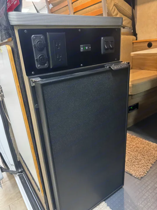 RV rental utility panel: integrated refrigerator, AC outlets, 12V sockets, and a "Coach Batt" battery monitor.. Winnebago Solis 2025