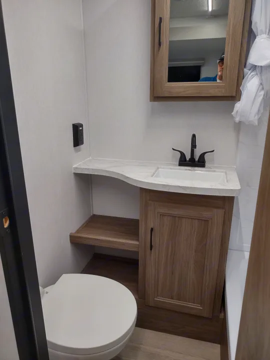 Compact modern RV rental bathroom featuring a wood vanity, sink, mirror, toilet, open shelf, and black faucet.. Coachmen 29 SE 2025