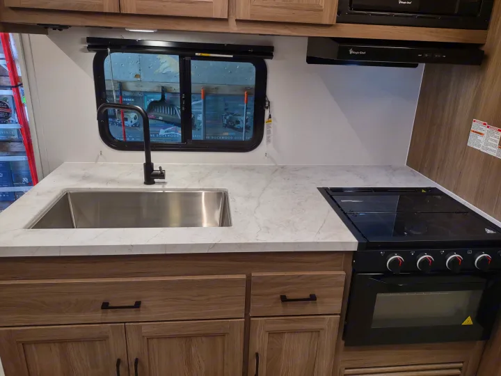RV rental kitchen interior: stainless sink, black faucet, stovetop/oven, light wood cabinets, and marble-look countertop.. Coachmen 29 SE 2025