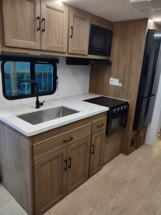 Modern RV rental kitchen with light wood cabinetry, white countertops, stainless sink, black appliances & a window.. Coachmen 29 SE 2025
