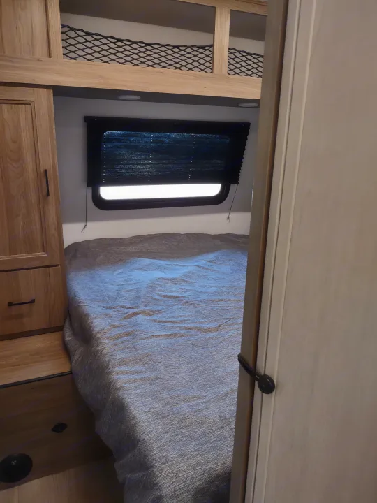 RV rental bedroom: a cozy bed, window with blind, wooden cabinet, and an upper bunk with safety netting, seen from a doorway.. Coachmen 29 SE 2025