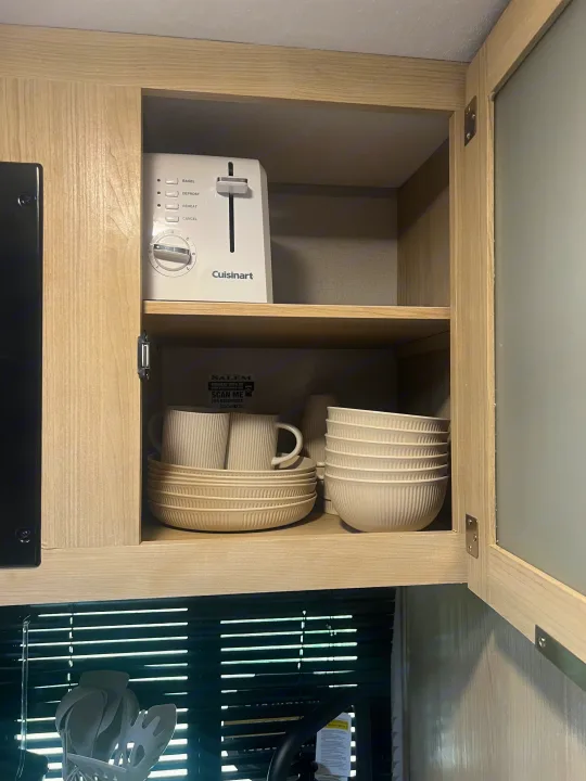 RV rental kitchen cabinet with a Cuisinart toaster on the top shelf and stacked light-colored dishes below.. Forest River Salem FSX 2023