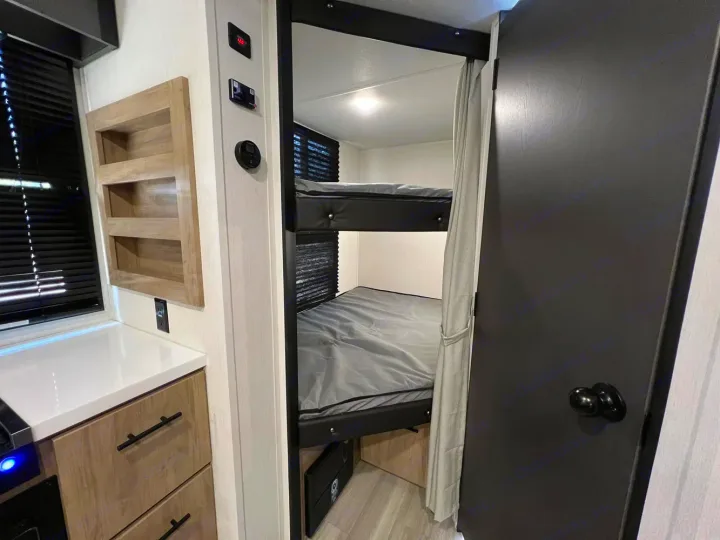 Inside an RV rental, a compact bunk bed area with gray bedding is visible. To the left, a counter, storage, and wall controls are shown.. Forest River Campsite Reserve 2026