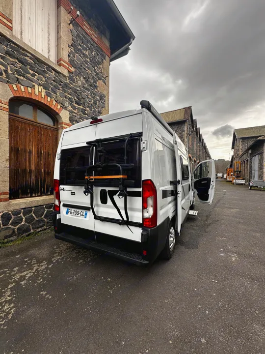 White camper van (RV rental) with a bike rack, parked by an old stone building under a cloudy sky, ready for adventure.. mc louis menfys 2023