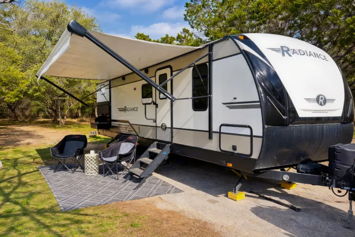 Radiance RV rental with an extended awning, two chairs, and a rug on a sunny campsite. The travel trailer is white and black.. Cruiser RV Radiance Ultra Lite 2020
