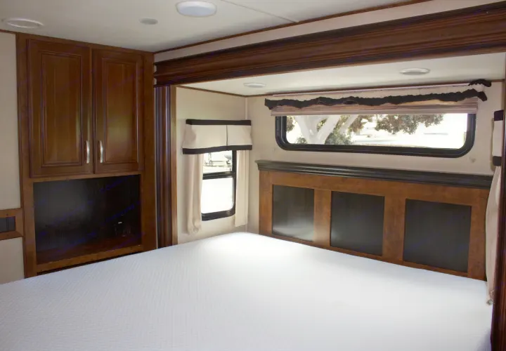 Inside an RV rental bedroom, featuring a pristine bed, dark wood cabinets, and two windows for natural light.. Grand Design Solitude 2015