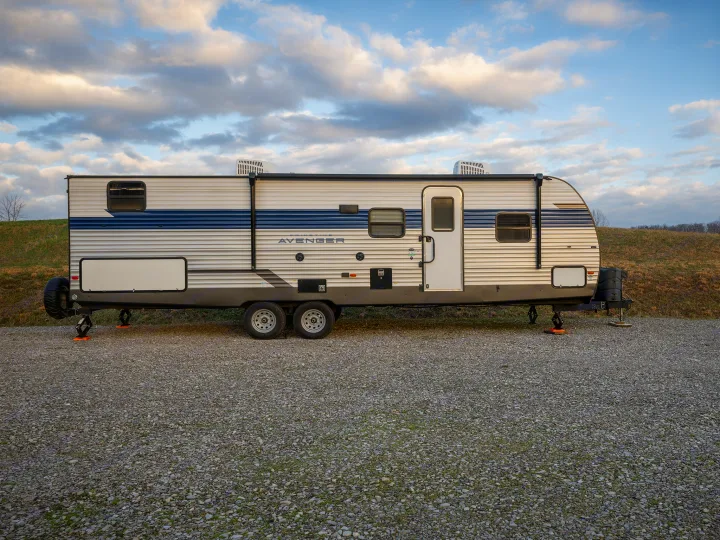 An Avenger travel trailer rv rental, parked on gravel under a cloudy sky, awaits its next adventure or camping trip.. Prime Time Avenger 2024