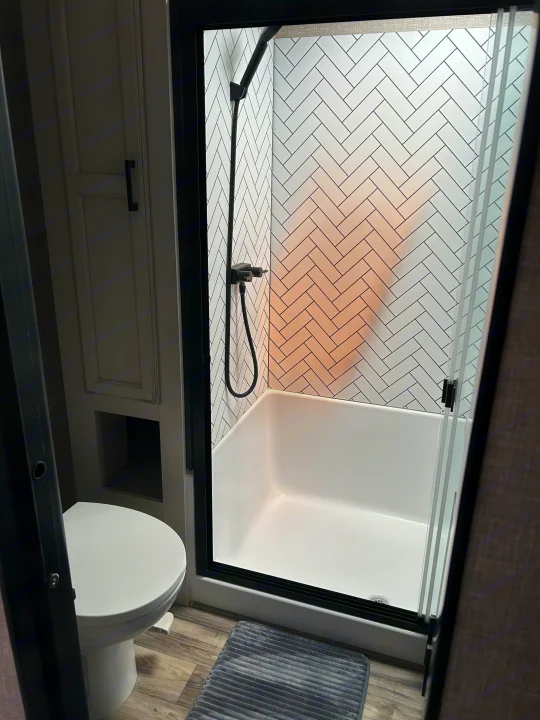 RV rental bathroom with modern shower, white herringbone tile, and toilet. A gray bath mat is on the wood-look floor.. Jayco Eagle 2022