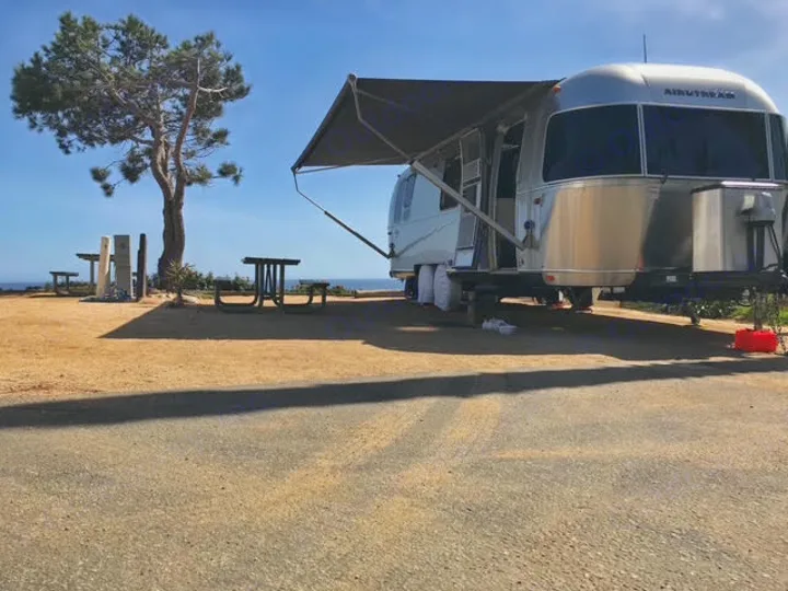 This RV rental, a shiny Airstream, enjoys a sunny campsite with picnic tables, a large tree, and an ocean view.. Airstream International 2016