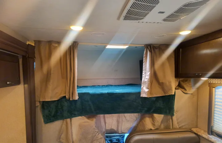 Overhead bunk bed with teal blanket & beige privacy curtains in an RV rental interior, featuring ceiling lights & AC vents.. Thor Motor Coach Freedom Elite 2019