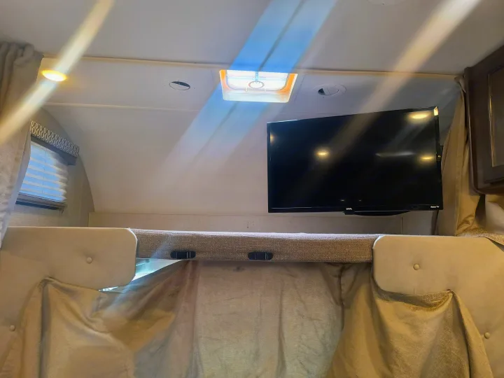 RV rental interior showing a cozy bunk bed with a safety net, mounted TV, ceiling lights, and window with blinds.. Thor Motor Coach Freedom Elite 2019