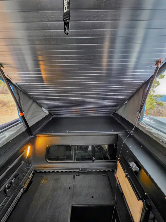 Explore this spacious rv rental's pop-top camper interior, featuring a raised sleeping area, side windows, and convenient storage.. Dirtbox Overland Canopy Camper 2023