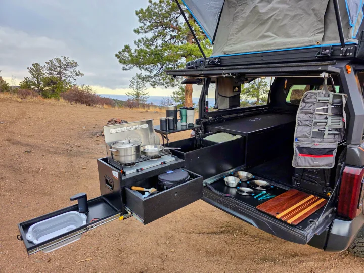RV rental truck's pull-out kitchen with stove, sink, and outdoor cooking gear. Rooftop tent deployed for camping.. Dirtbox Overland Canopy Camper 2023