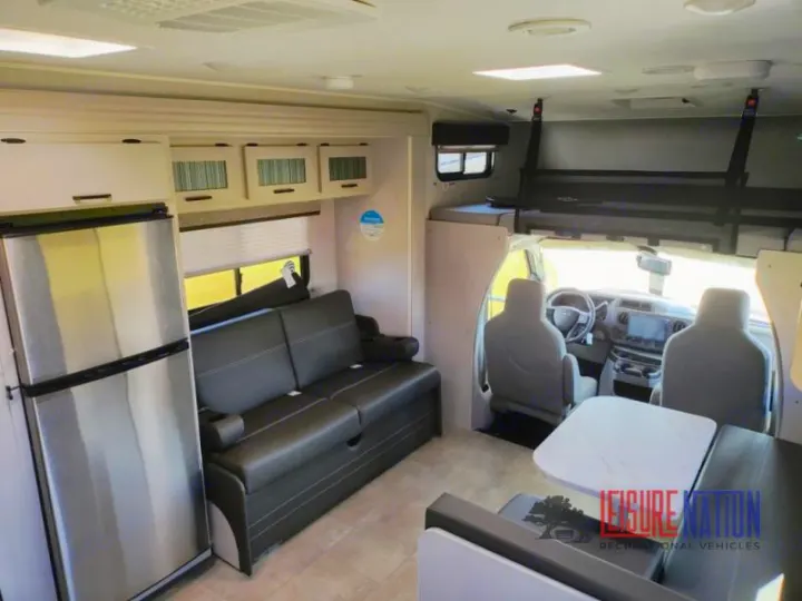 Spacious rv rental interior featuring a refrigerator, sofa, dinette, and an overhead bunk bed above the cab area.. Coachmen Freelander 2023