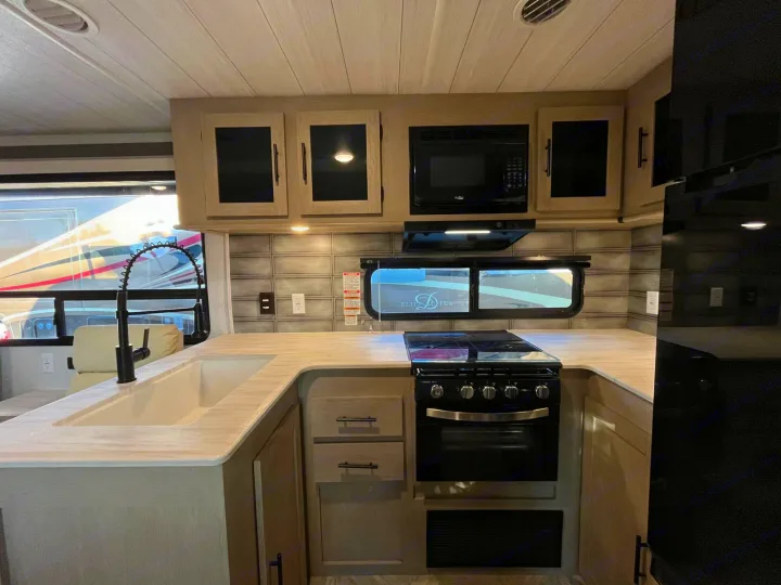 Modern RV rental kitchen: light wood cabinets, black appliances, gooseneck faucet, sink, and outside views through windows.. Forest River Salem 2023