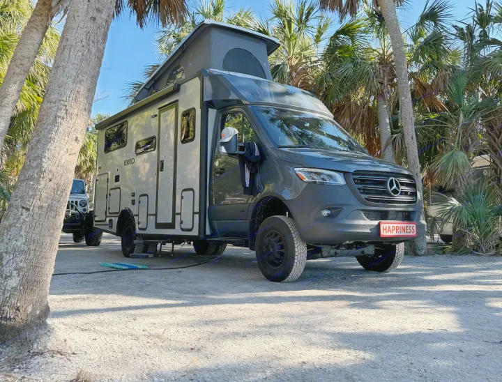 Gray EKKO RV rental (Mercedes camper van) with pop-top, parked on a shell lot under palm trees. Its plate reads HAPPINESS!. Winnebago Ekko 2025
