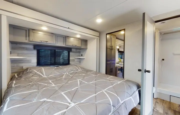 This modern RV rental bedroom features a comfy bed, overhead storage, and window, offering a glimpse of the shower and dining area.. Crossroads RV Volante 2023