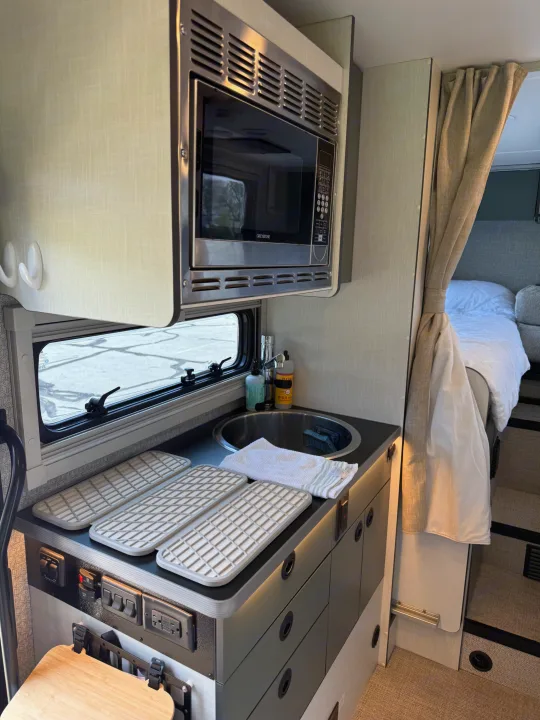 Inside an RV rental: modern kitchen with microwave, sink, drying mats, controls, and a peek into the cozy bed space.. Winnebago Ekko 2025