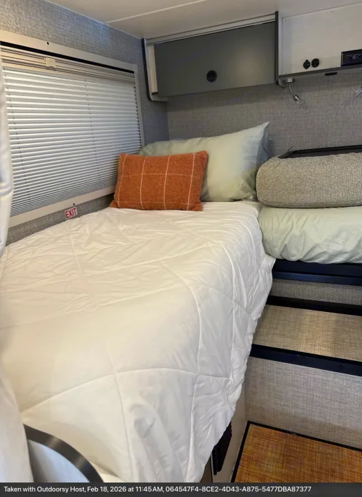 Cozy RV rental interior featuring a comfortable bed with white bedding, pillows, window blinds, and overhead storage.. Winnebago Ekko 2025