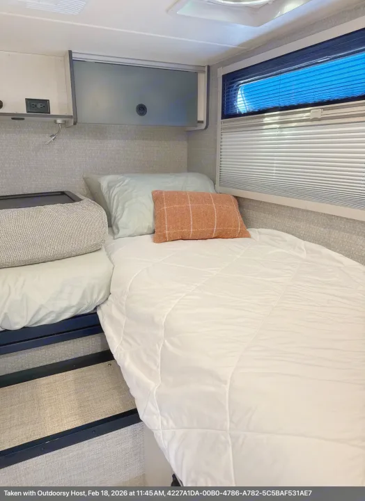 Cozy RV rental bedroom featuring a comfortable bed with soft bedding, pillows, and a window with blinds showing bright blue sky.. Winnebago Ekko 2025