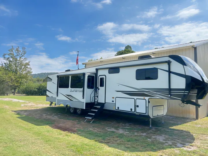 A white Durango fifth-wheel RV rental, with an American flag on its roof, is parked on grass next to a metal building.. Durango Durango Fifth Wheel 2023
