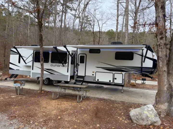 Spacious white fifth-wheel RV rental at a wooded campsite with extended awnings, picnic table, and grill for camping.. Durango Durango Fifth Wheel 2023