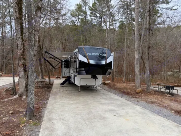 A modern Durango fifth-wheel rv rental is set up at a wooded campsite, featuring extended awnings & picnic tables.. Durango Durango Fifth Wheel 2023