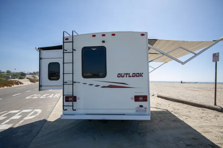 Outlook RV rental with extended awning by a sandy beach. The white vehicle is parked near a road with the ocean beyond.. Winnebago Outlook 2019