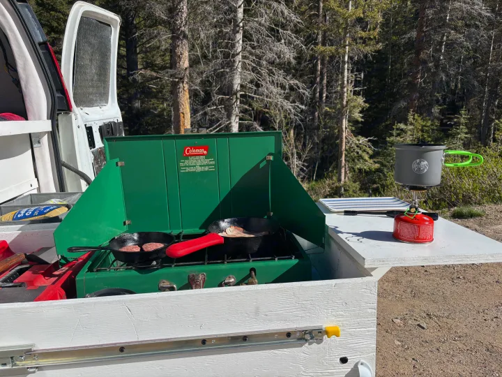 RV rental outdoor kitchen in a forest. Cooking breakfast on a Coleman stove with patties & bacon, plus a portable stove for hot drinks.. Chevrolet Express 1500 AWD 2007