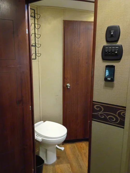 Inside an RV rental: View of the compact bathroom with a white toilet, wooden door, and control switches on the wall.. Keystone RV Outback Super-Lite 2015