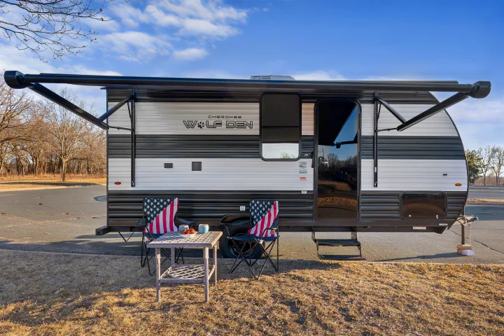 Cherokee Wolf Den travel trailer RV rental parked outdoors, featuring an extended awning, American flag chairs, and a table with strawberries.. Forest River Wolf Den 16EV 2025