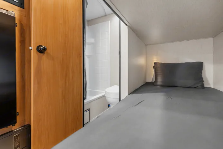Cozy RV rental interior with a dark-sheeted bunk bed on the right, and a compact bathroom with toilet and shower on the left.. Forest River Wolf Den 16EV 2025