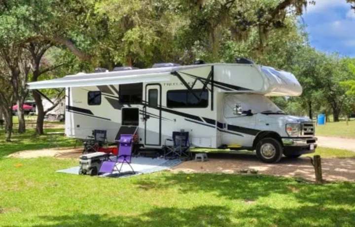 Enjoy this rv rental motorhome setup at a shaded campsite: awning extended, chairs, and cooler ready for outdoor relaxation.. Coachmen Freelander 2021