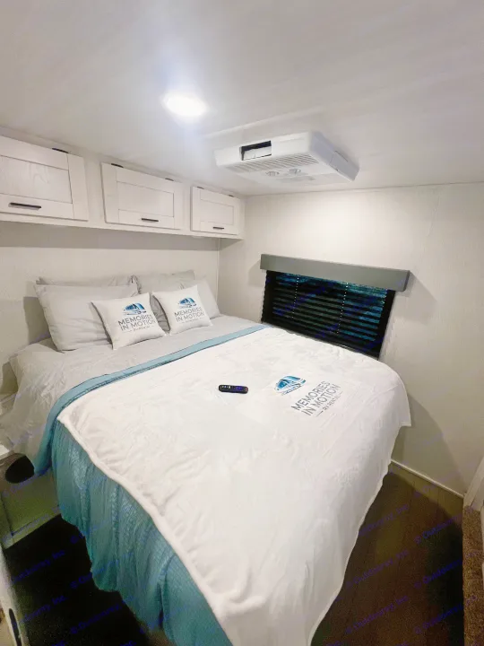 Cozy RV bedroom with a bed featuring "Memories in Motion" branded linens, overhead cabinets, and AC unit for your RV rental.. 1FDXE4FN9RDD14726 3100FB 2023