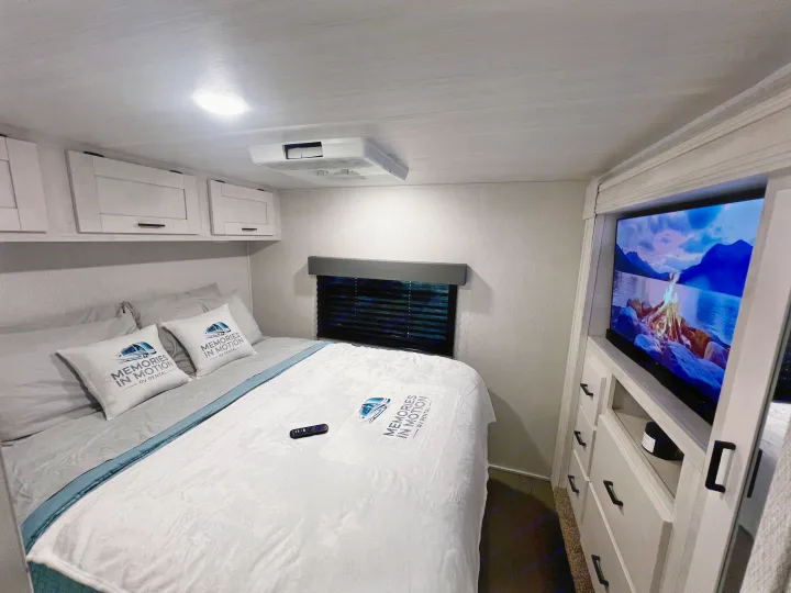 Cozy RV rental bedroom with "Memories In Motion" branded bedding, overhead cabinets, a mounted TV, and window blinds.. 1FDXE4FN9RDD14726 3100FB 2023